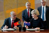 Agio Pereira and Julie Bishop sign paperwork in front of small East Timor and Australian flags.