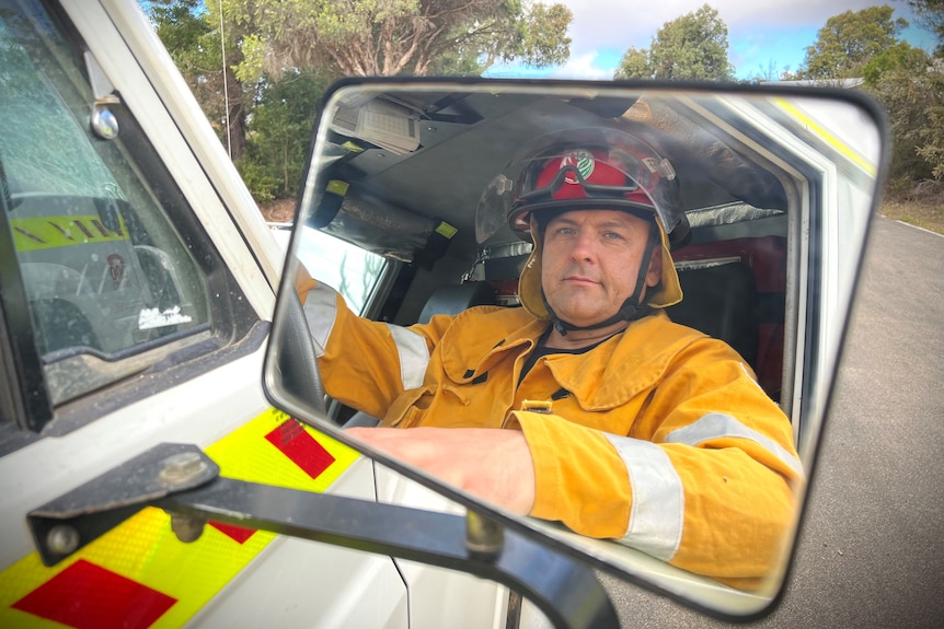 Male firefighter looks in side mirror in car wearing red helmet and yellow protective uniform with hand out the window. 