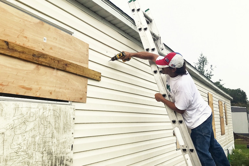 A man reinforces drills boards over windows as he reinforces his mobile home.