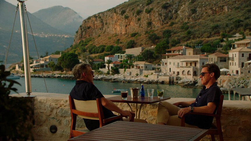 Two men in shirts, shorts and sunglasses sit and talk at outdoor dining table with view of Greek seaside town at sunset.