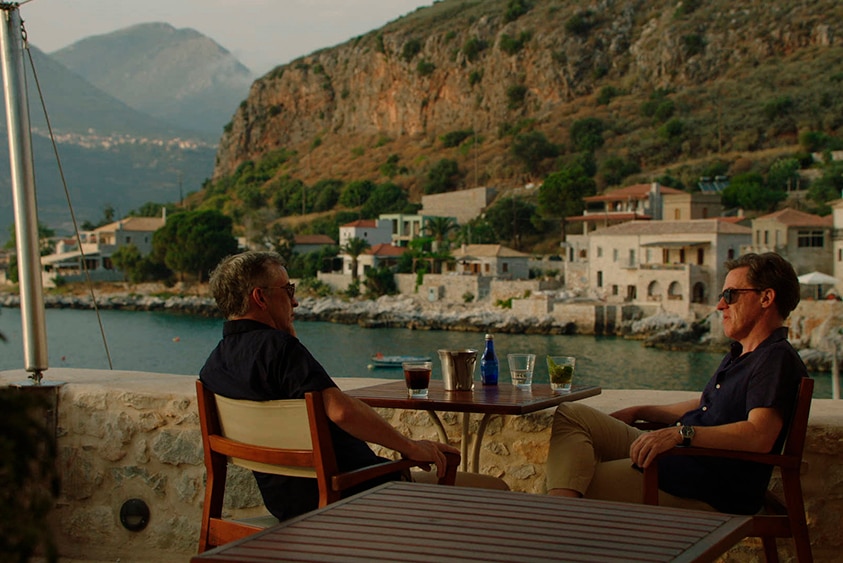 Two men in shirts, shorts and sunglasses sit and talk at outdoor dining table with view of Greek seaside town at sunset.