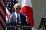 President Biden wears sunglasses in a suit speaks behind a lectern to a journalist.