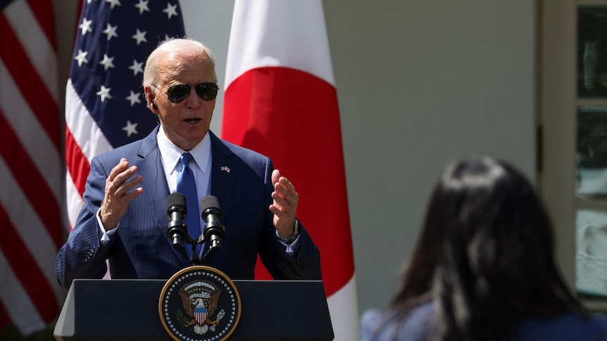 President Biden wears sunglasses in a suit speaks behind a lectern to a journalist.