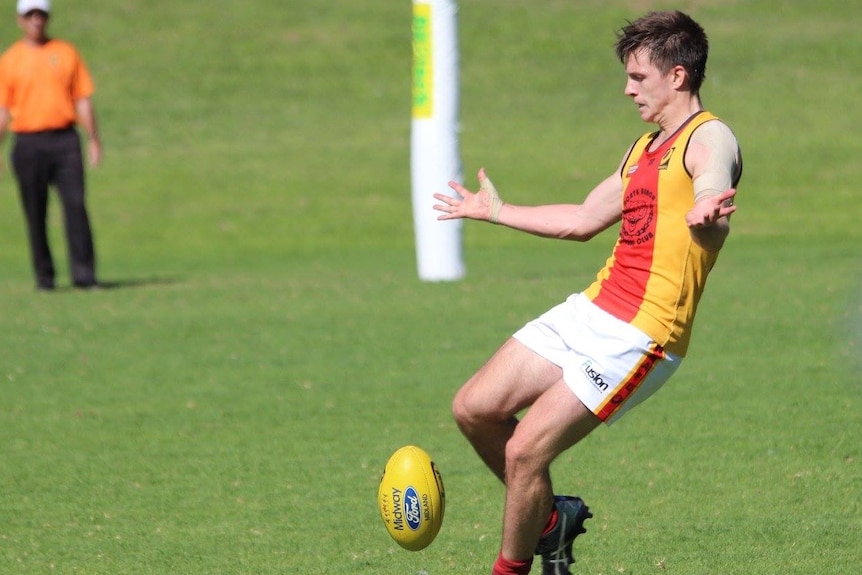 Football player in the motion of kicking. Green grass surrounds him with a goal umpire watching on in the background.