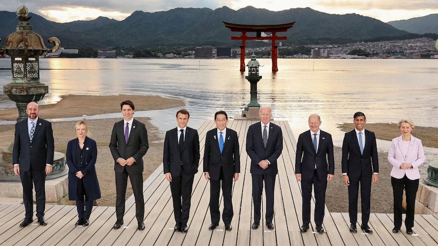 G7 leaders line up for photo call by the seaside, with a Japanese mon (gate) seen in the water behind.