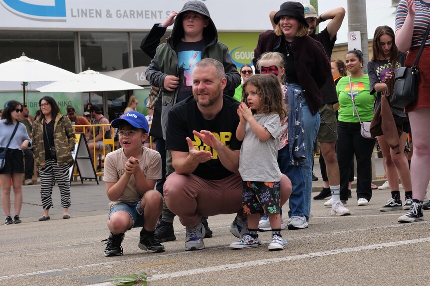 A man crouches down and claps with two small children standing beside him watching the stage.