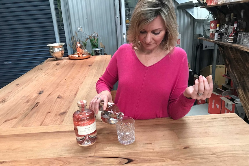 A woman pours a nip of gin from a bottle into a crystal tumbler at a wooden bar inside a large shed.