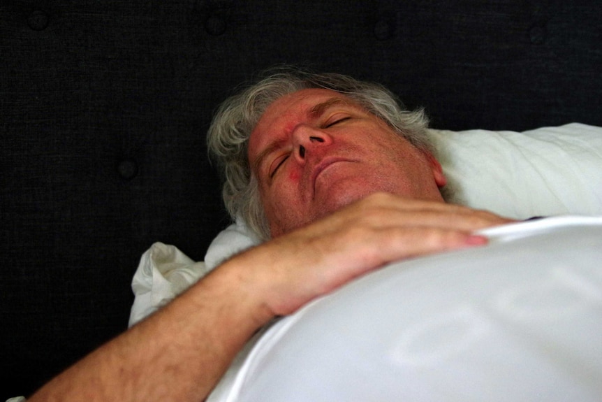 A close-up shot of a man with grey hair lying down on his back with a hand on his chest.