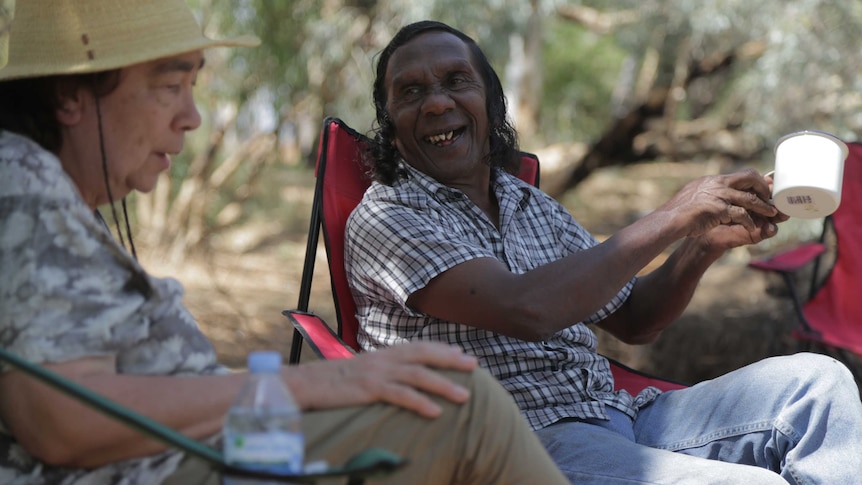 A man sitting in a folding chair holds a mug in his hand as he talks with a woman wearing a hat.