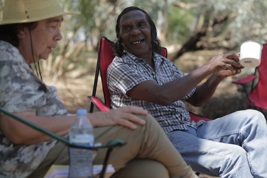 A man sitting in a folding chair holds a mug in his hand as he talks with a woman wearing a hat.