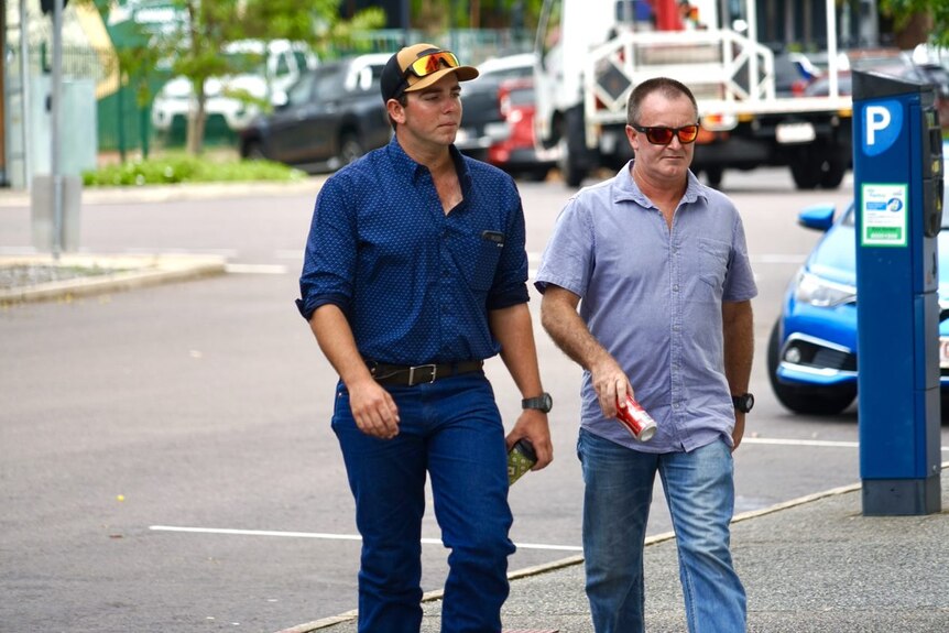 Two men crossing a road, the younger one wearing a navy shirt, jeans and sunglasses on top of his cap.