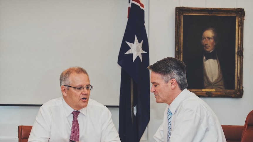 Morrison (right) and Cormann (left) sit talking at a table covered in documents with an Australian flag in the background