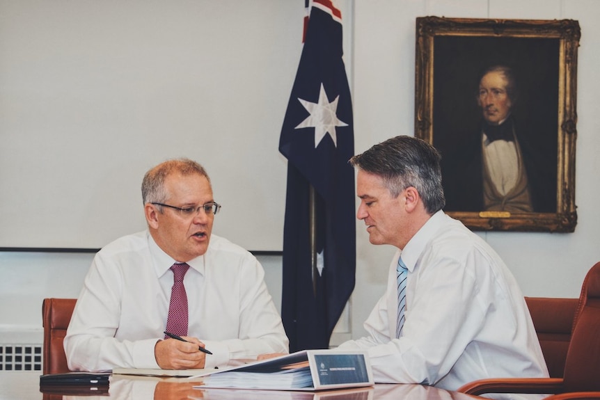 Morrison (right) and Cormann (left) sit talking at a table covered in documents with an Australian flag in the background