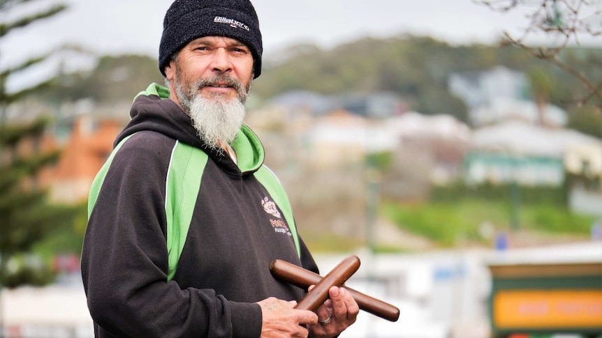 An older Indigenous man with a long grey beard, wearing a hoodie and holding clapsticks.