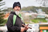 An Indigenous man standing outside wearing a beanie and hoodie, holding a pair of clapsticks.