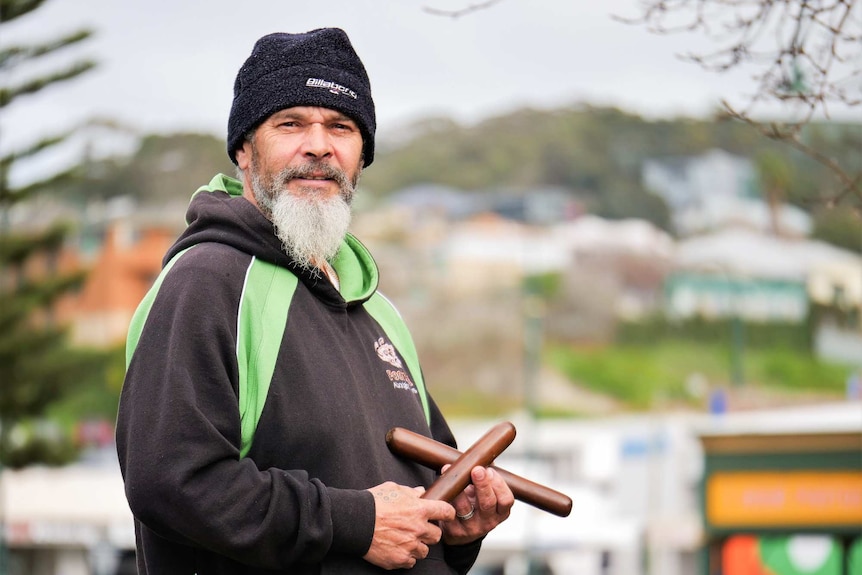 An Indigenous man wearing a beanie and a hoodie, standing outside and holding clapsticks.