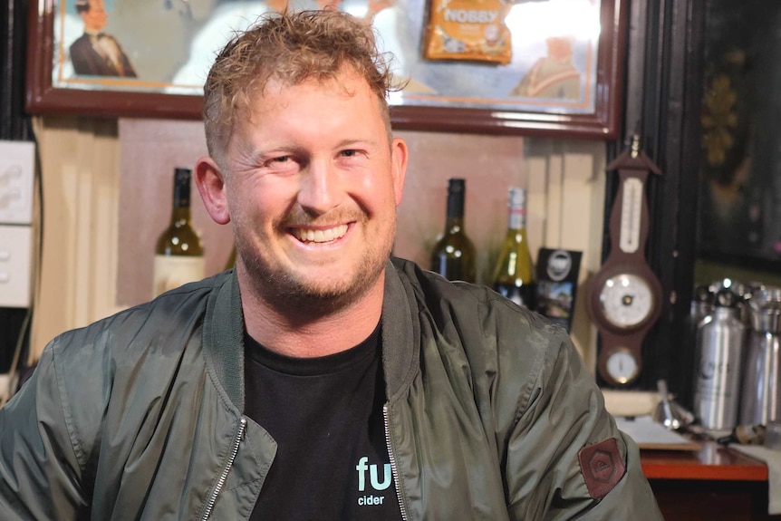 A man with fair hair and a big smile sits at a bar with wine bottles behind him.