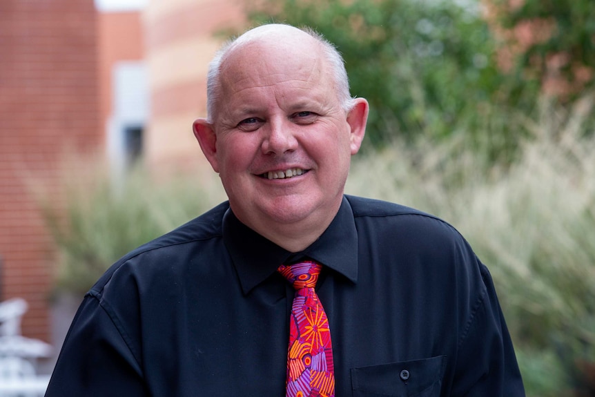 A white-haired man wearing a colourful tie smiles for the camera.