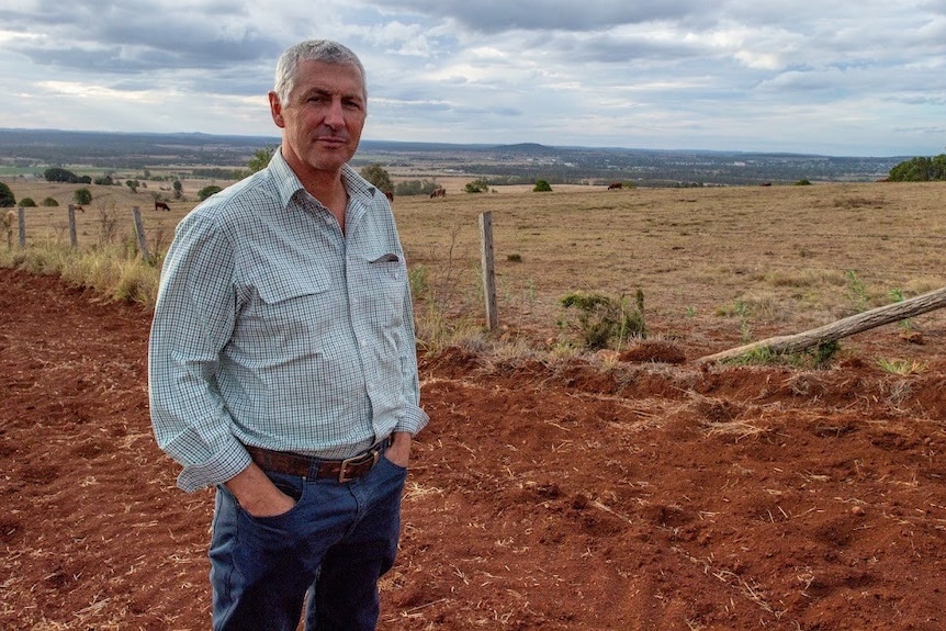 Un hombre de unos 60 años con cabello blanco corto, vestido con una camisa azul y jeans, parado en un campo de tierra roja.