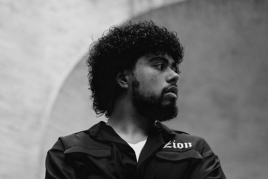 A black and white photo of a young man with thick, dark curly hair and a beard. 