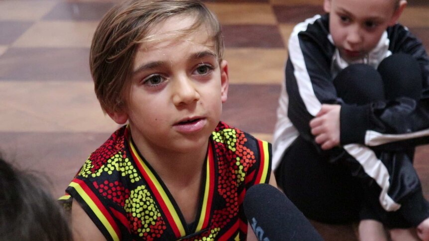 Young indigenous boy wearing an indigenous jersey, sitting on the floor talking into a microphone. 