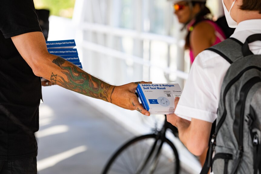 A man with a tattooed arm hands a rapid antigen test to a person.