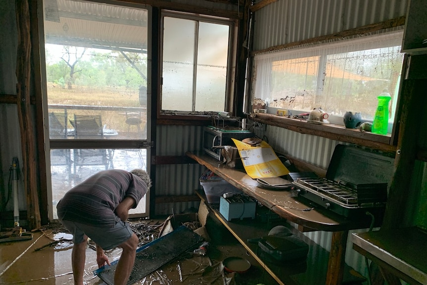 Man picks up items covered in mud after floodwaters, windows muddy.