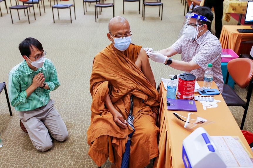 A Buddhist monk in golden robes gets his vaccine from a nurse while a man kneels in prayer beside him