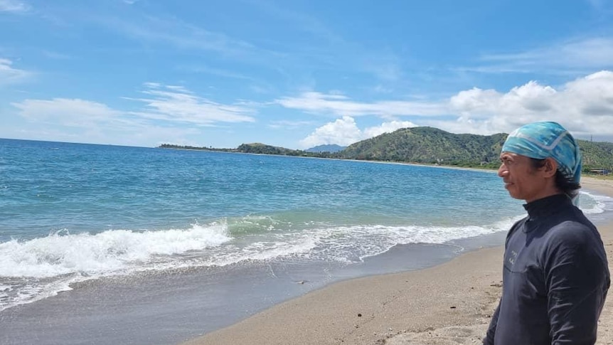 A man wearing a bandanna stands on the sand looking out to aqua blue ocean