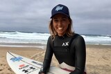 A female surfer sits on the beach with her surfboard and smiles for the camera.