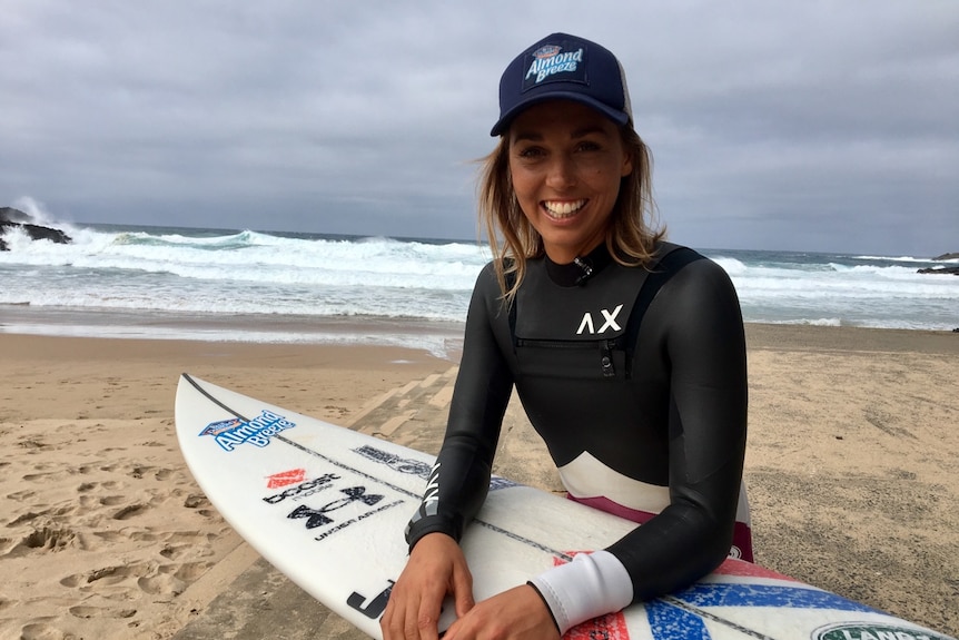 A female surfer sits on the beach with her surfboard and smiles for the camera.