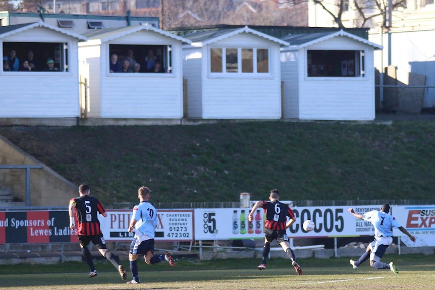 Four white beach huts sit atop a hill and people inside watch a soccer game below.