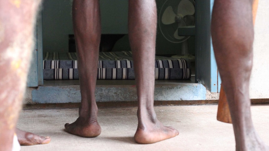 Groups of feet stand outside a room with a mattress on the floor in a prison block.