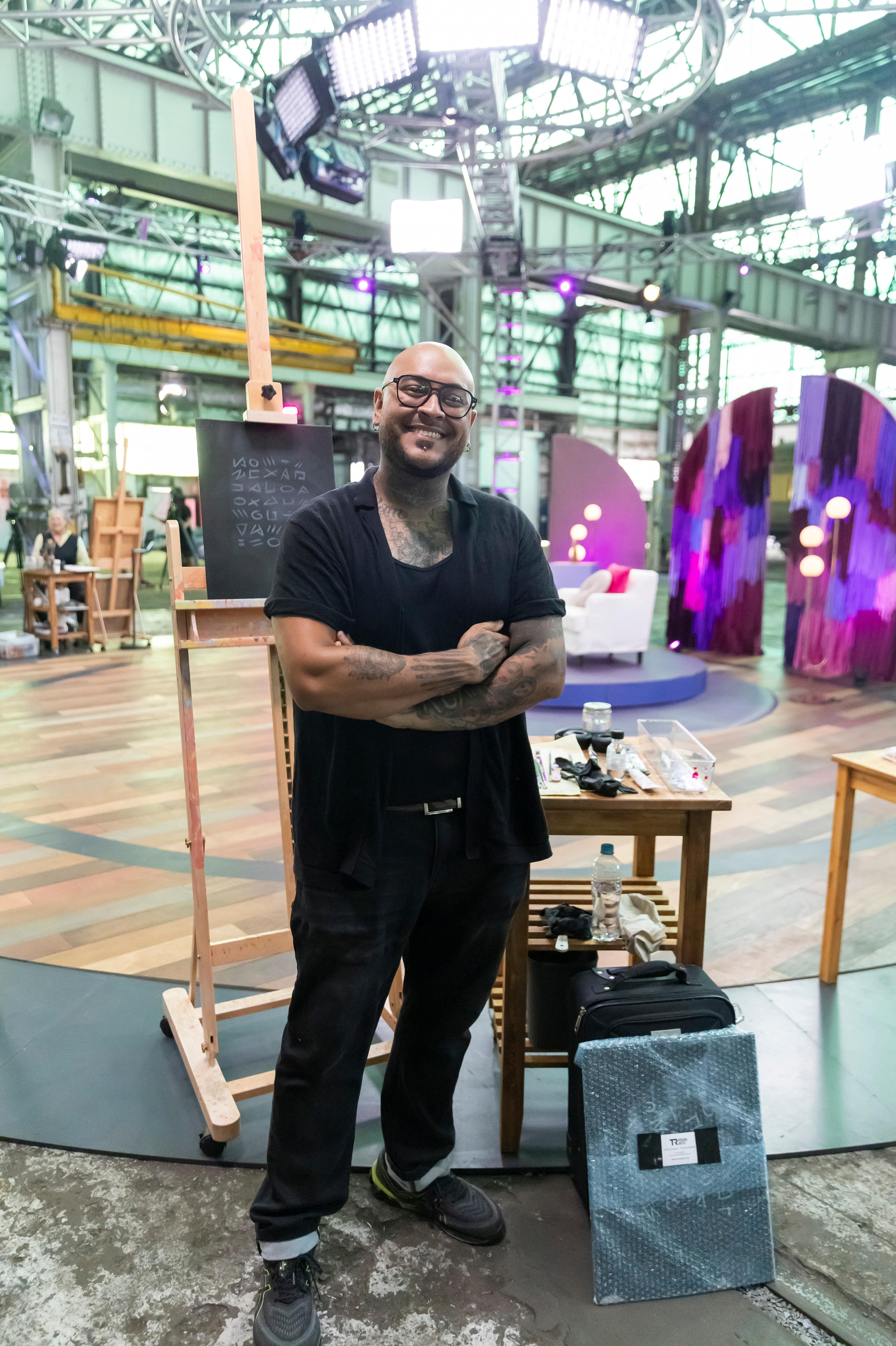A man with brown skin stands smiling at the camera with his arms folded in front of him. An easel and art supplies behind him