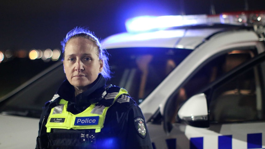 A police officer with her hair tied back, wearing a uniform, looks at the camera at night, with a police car behind her.