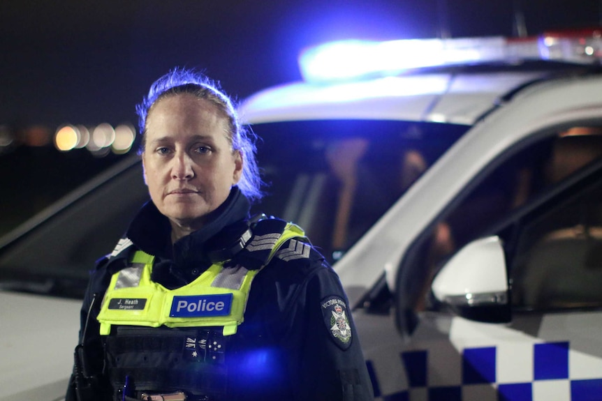 A police officer with her hair tied back, wearing a uniform, looks at the camera at night, with a police car behind her.