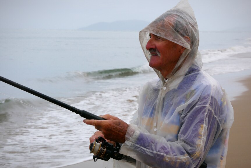A man wearing a rain poncho fishes on the edge of a beach in the rain.