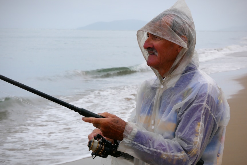 Man in raincoat fishing at the edge of the beach in the rain.