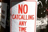 A red and white sign stuck on a pole says no catcalling any time.