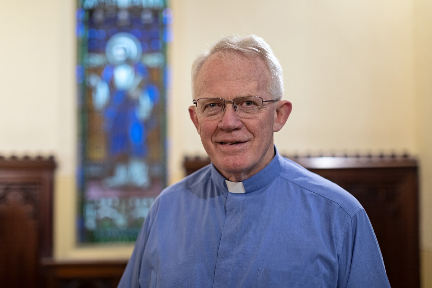 Rector Craig Segaert wearing blue shirt with white collar, with stained glass window behind him.