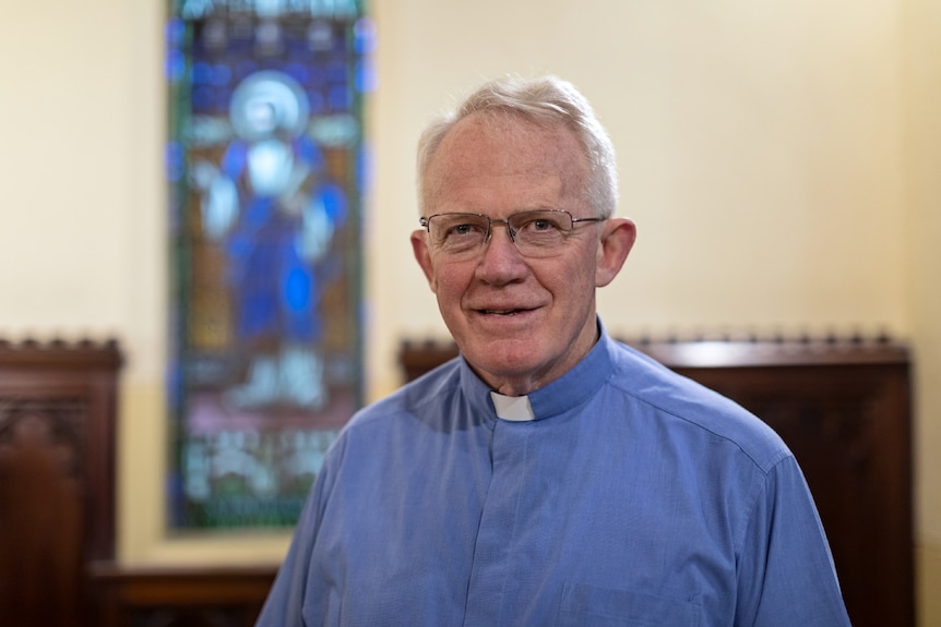 Rector Craig Segaert wearing blue shirt with white collar, with stained glass window behind him.