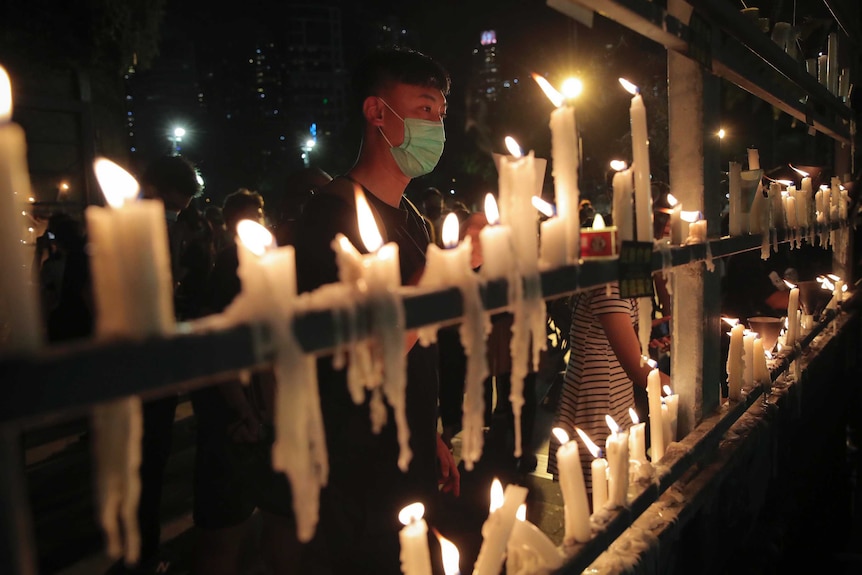 People light a long row of candles as crowds can be seen in the background on a dark night.