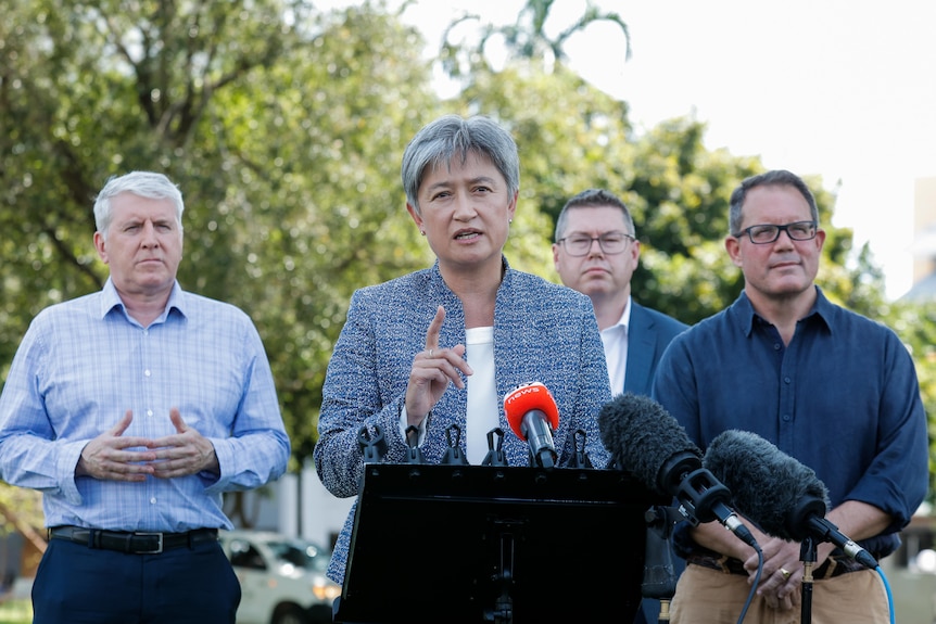 Penny Wong speaks at an outdoor media conference. Three men stand behind her. 