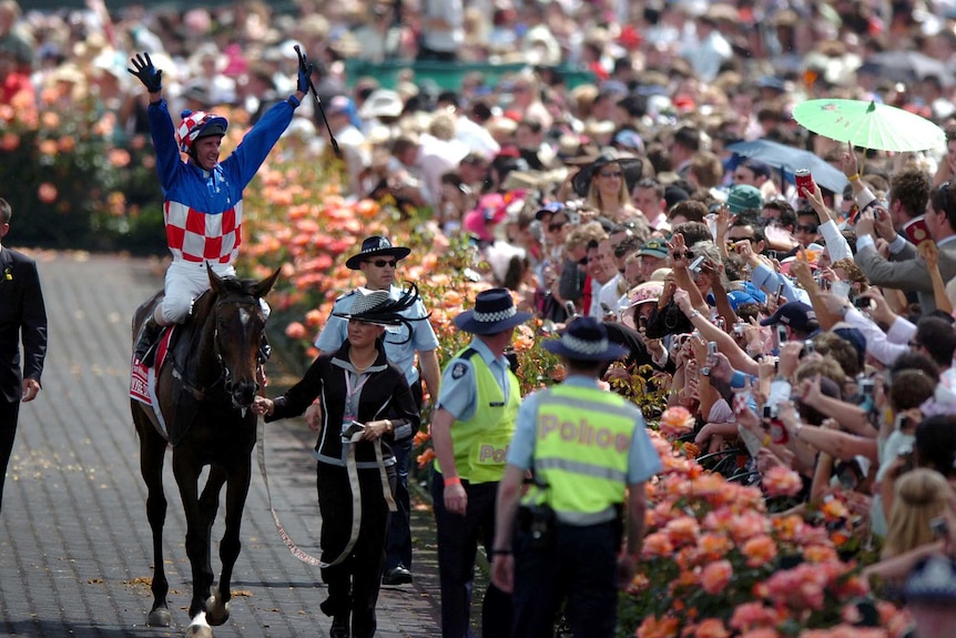 A jockey atop a horse holds his hands high in the air and waves to the crowd at a busy racetrack