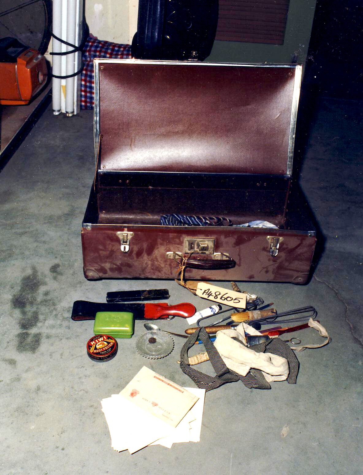 An open red suitcase with a white tag with numbers, its contents, including boot polish, strewn on the floor.