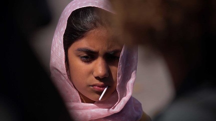 A young Muslim girl with a headscarf around her head and a lollypop in her mouth