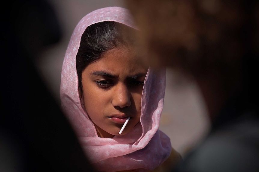 A young Muslim girl with a headscarf around her head and a lollypop in her mouth