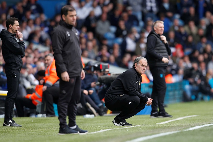 Marcelo Bielsa (centre) crouches down on the side of a football pitch at Elland Road.