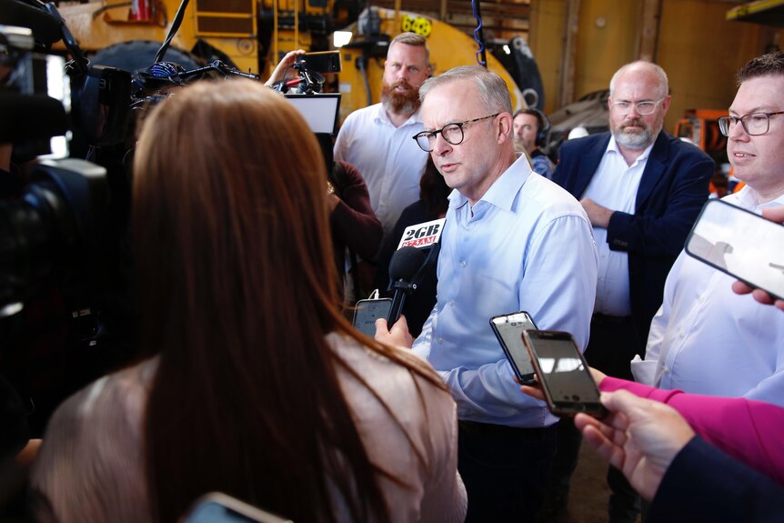 A man wearing glasses speaks while surrounded by a group of media workers