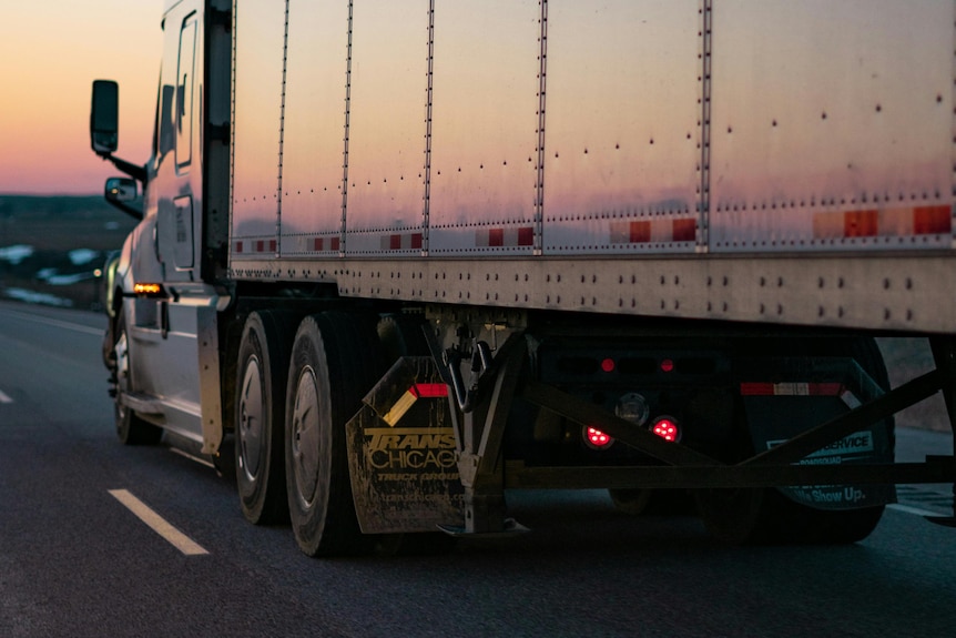 A large silver truck drives off into a pink sunset.